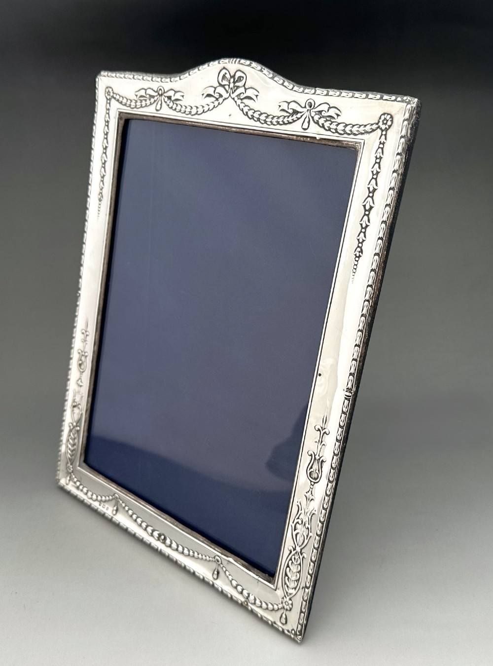 An Edwardian sterling silver portrait photo frame featuring embossed ribbon, garland, and swag decorative motifs with an arched top. View II