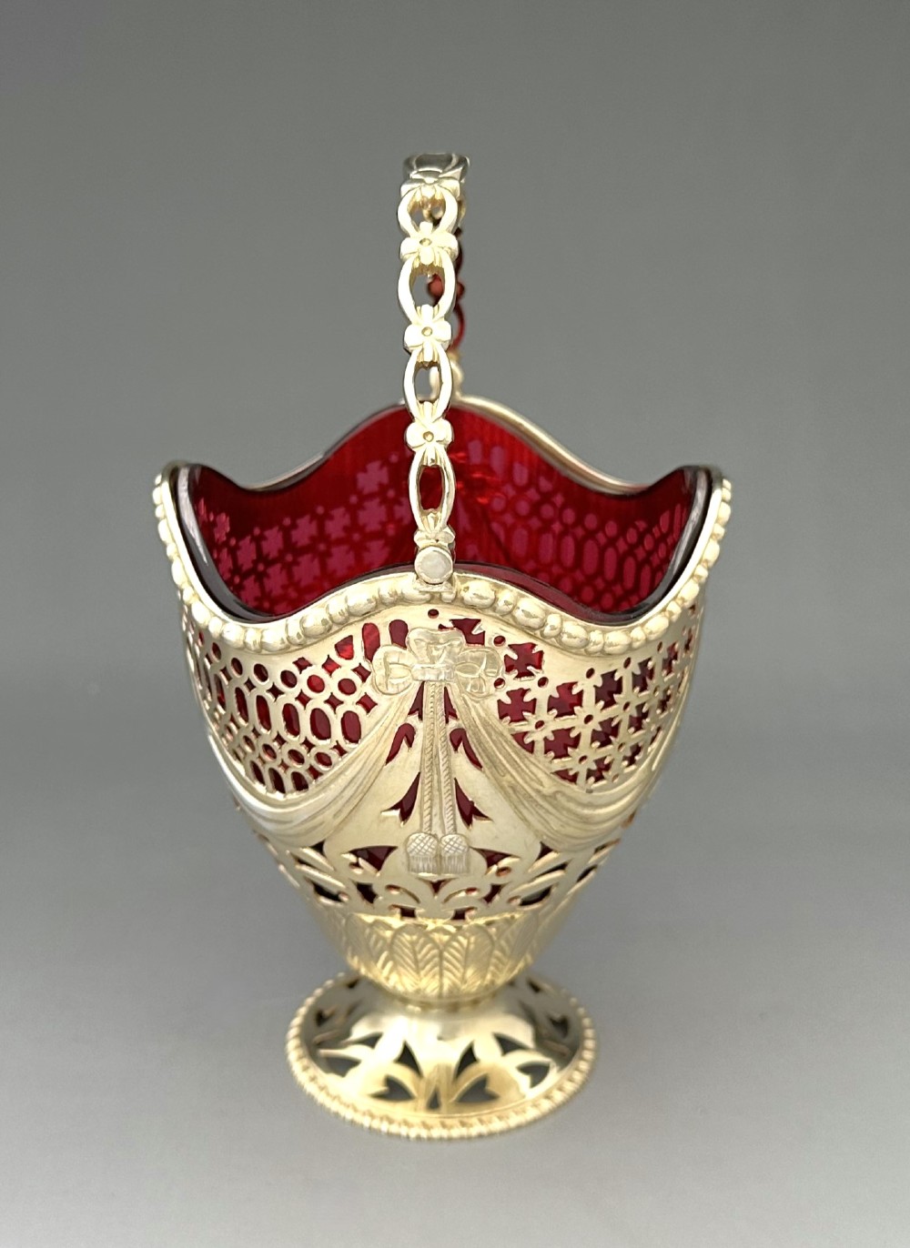 A pierced silver-gilt sugar basket featuring drape and bow motifs with an overhead swing handle, housing a scalloped ruby red cranberry glass liner. View II