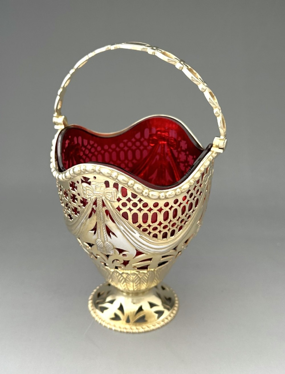 A pierced silver-gilt sugar basket featuring drape and bow motifs with an overhead swing handle, housing a scalloped ruby red cranberry glass liner. View III
