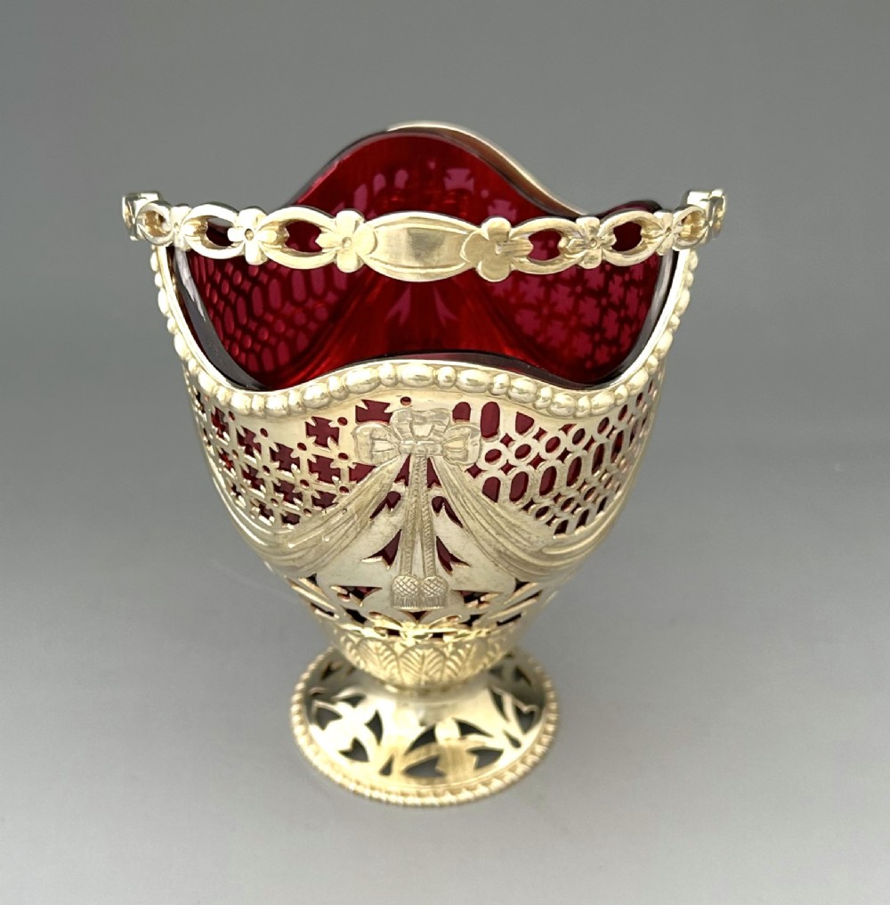 A pierced silver-gilt sugar basket featuring drape and bow motifs with an overhead swing handle, housing a scalloped ruby red cranberry glass liner. View IV