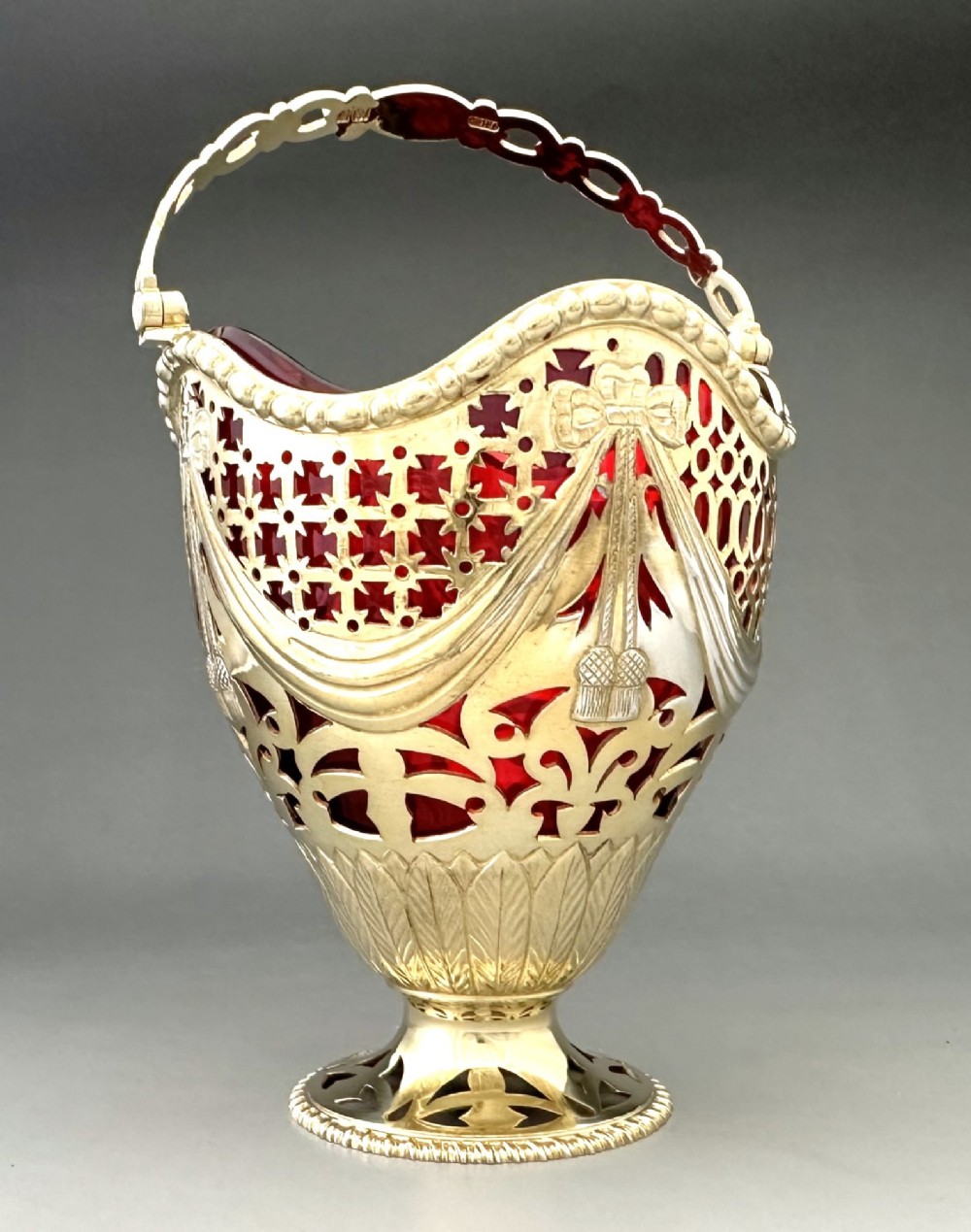 A pierced silver-gilt sugar basket featuring drape and bow motifs with an overhead swing handle, housing a scalloped ruby red cranberry glass liner. View V
