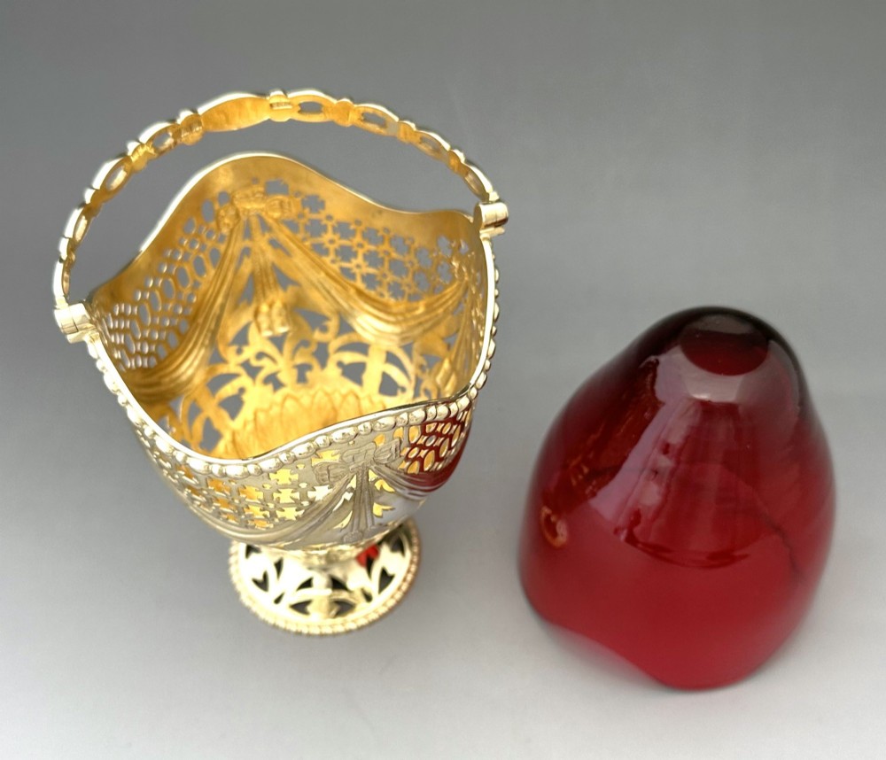 A pierced silver-gilt sugar basket featuring drape and bow motifs with an overhead swing handle, housing a scalloped ruby red cranberry glass liner. View VI