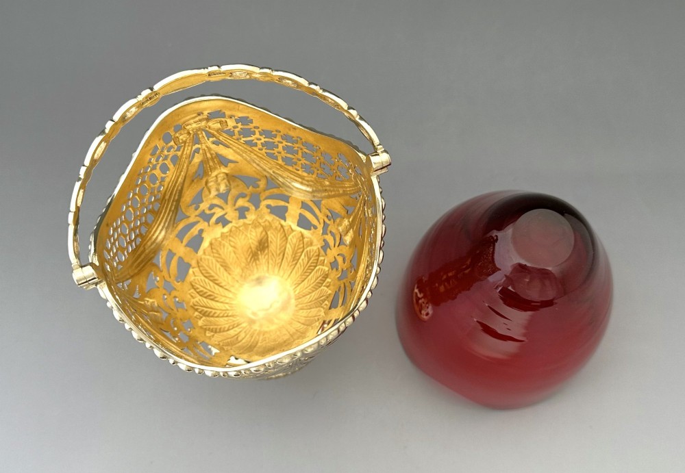 A pierced silver-gilt sugar basket featuring drape and bow motifs with an overhead swing handle, housing a scalloped ruby red cranberry glass liner. View VII