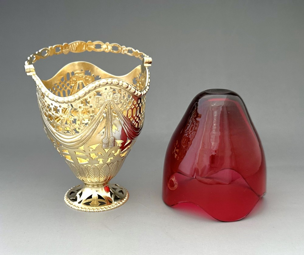 A pierced silver-gilt sugar basket featuring drape and bow motifs with an overhead swing handle, housing a scalloped ruby red cranberry glass liner. View VIII
