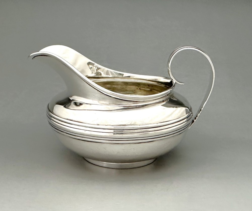 A George IV sterling silver cream jug with a polished surface, horizontal reeded decoration on the waist, a wide pour spout, and a curved high-loop handle.