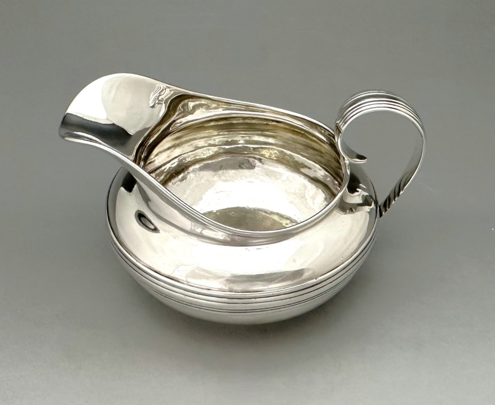 A George IV sterling silver cream jug with a polished surface, horizontal reeded decoration on the waist, a wide pour spout, and a curved high-loop handle. View II