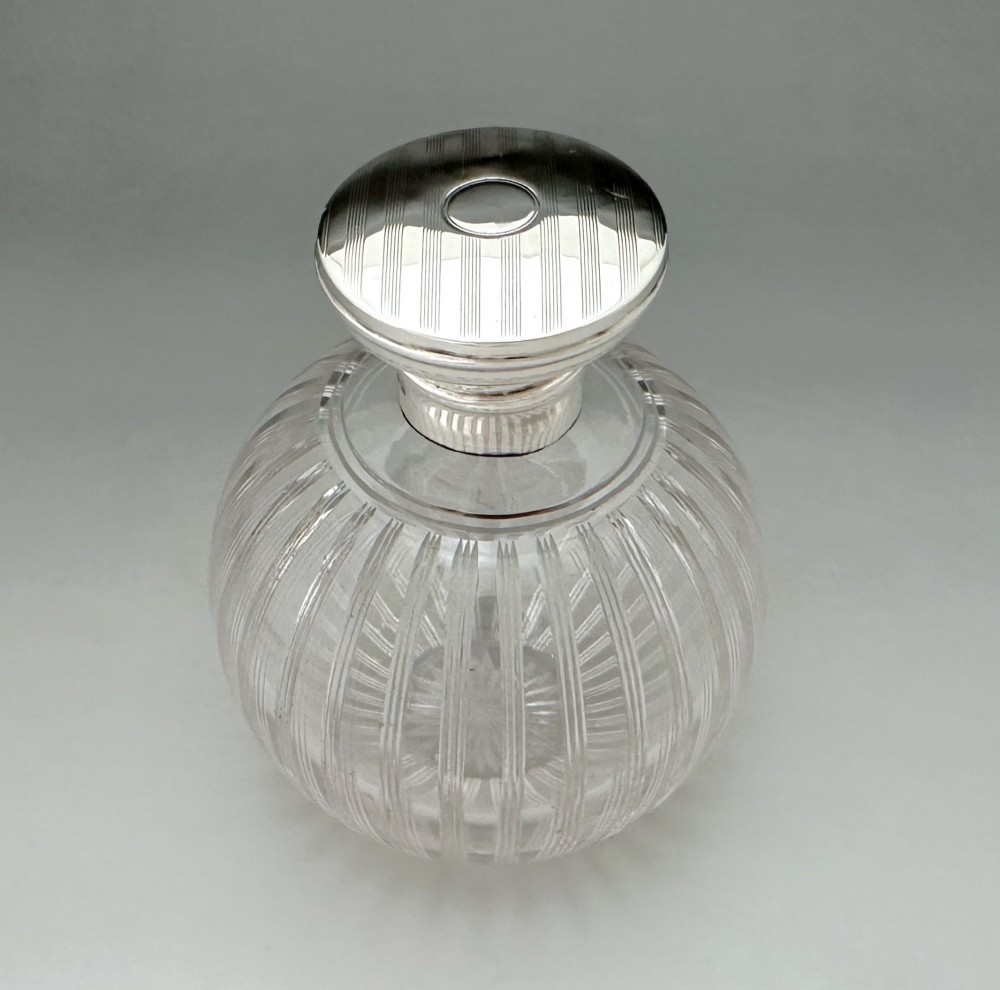 A globular clear fluted glass cologne bottle featuring a silver collar and an engine-turned circular top.