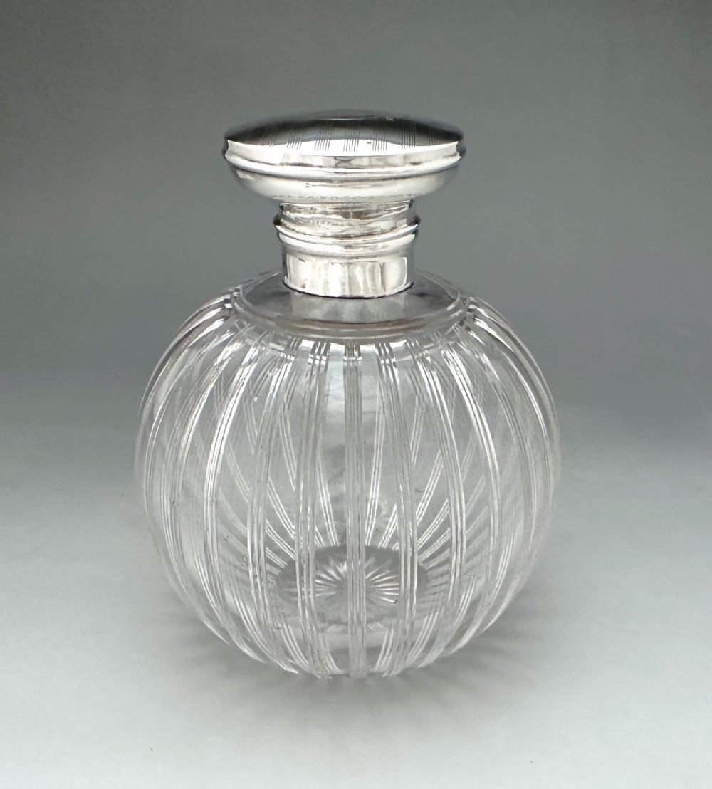 A globular clear fluted glass cologne bottle featuring a silver collar and an engine-turned circular top. View II