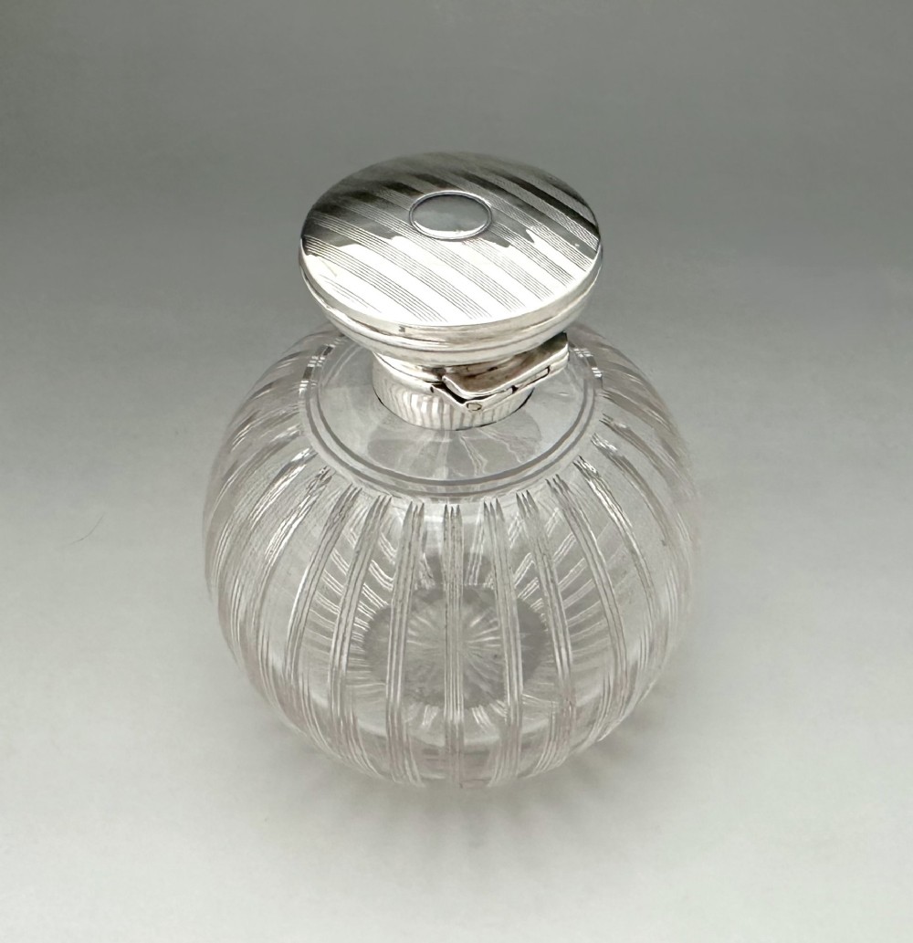A globular clear fluted glass cologne bottle featuring a silver collar and an engine-turned circular top. View III