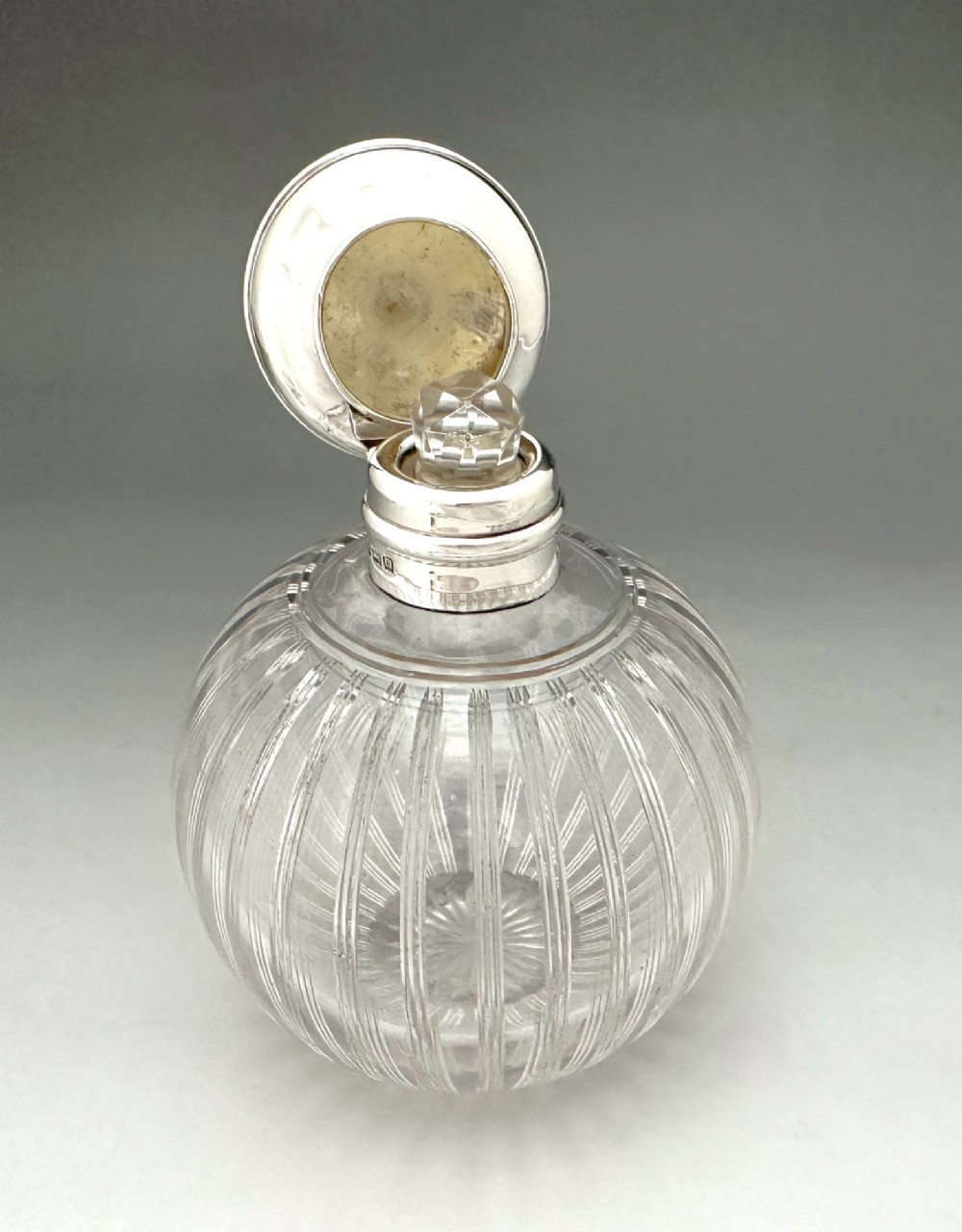 A globular clear fluted glass cologne bottle featuring a silver collar and an engine-turned circular top. View IV