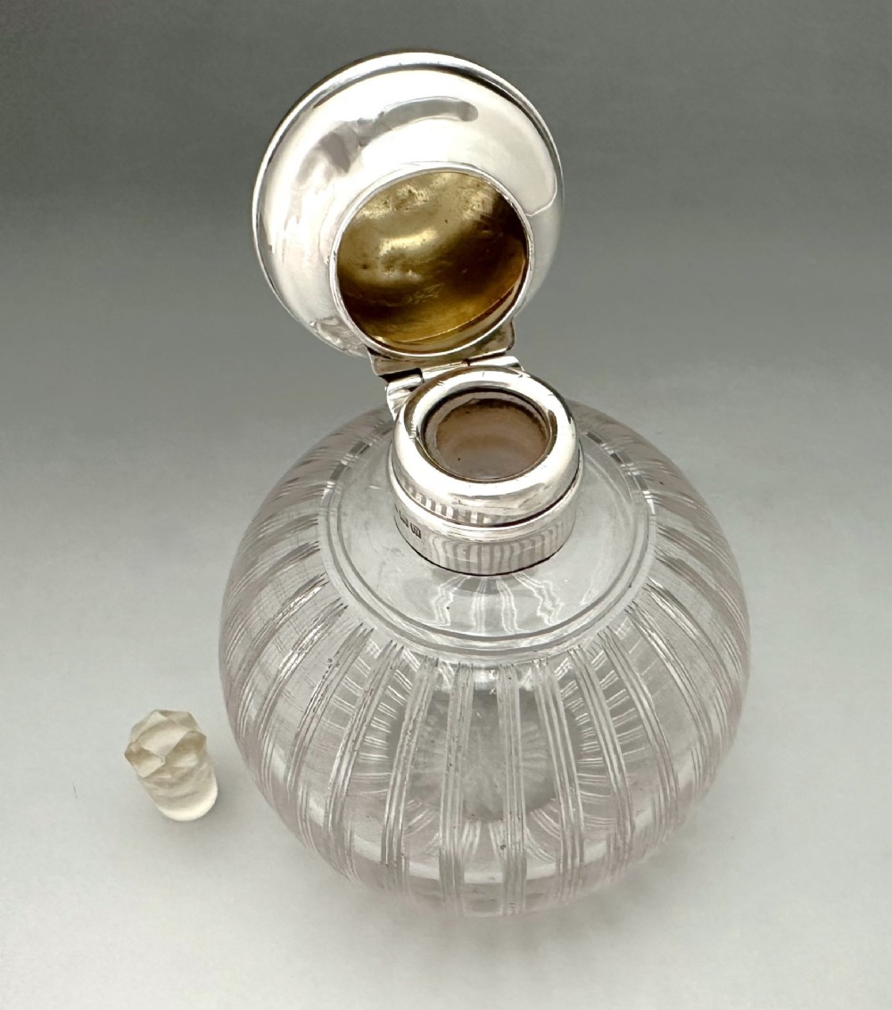 A globular clear fluted glass cologne bottle featuring a silver collar and an engine-turned circular top. View V
