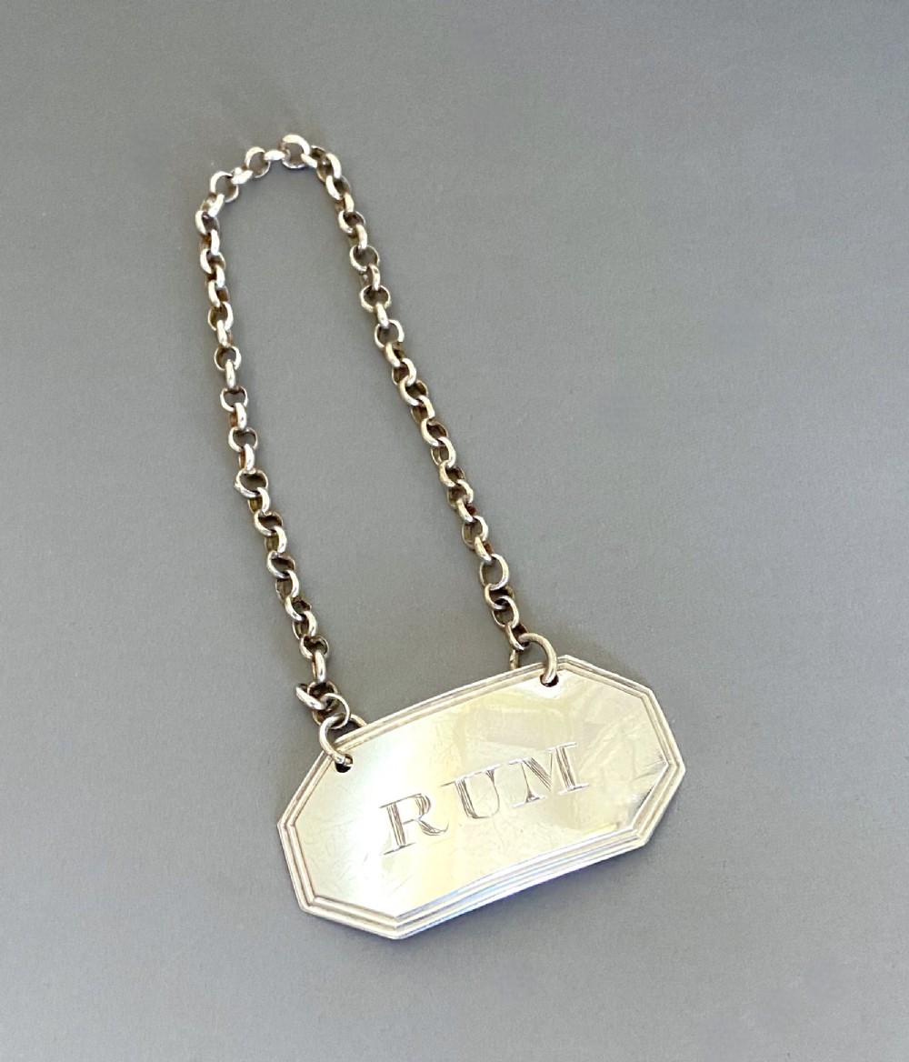 An octagonal sterling silver decanter label engraved with the word RUM, featuring a reeded border and suspended by a thin silver chain.