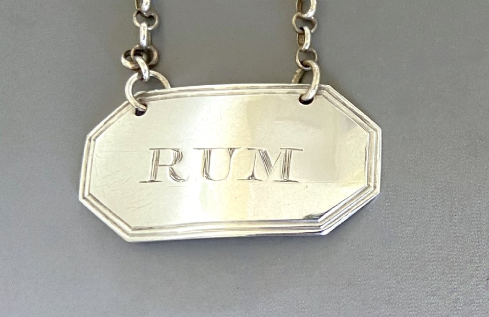 An octagonal sterling silver decanter label engraved with the word RUM, featuring a reeded border and suspended by a thin silver chain. View II