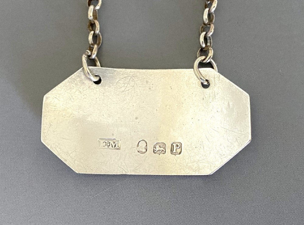 An octagonal sterling silver decanter label engraved with the word RUM, featuring a reeded border and suspended by a thin silver chain. View IV