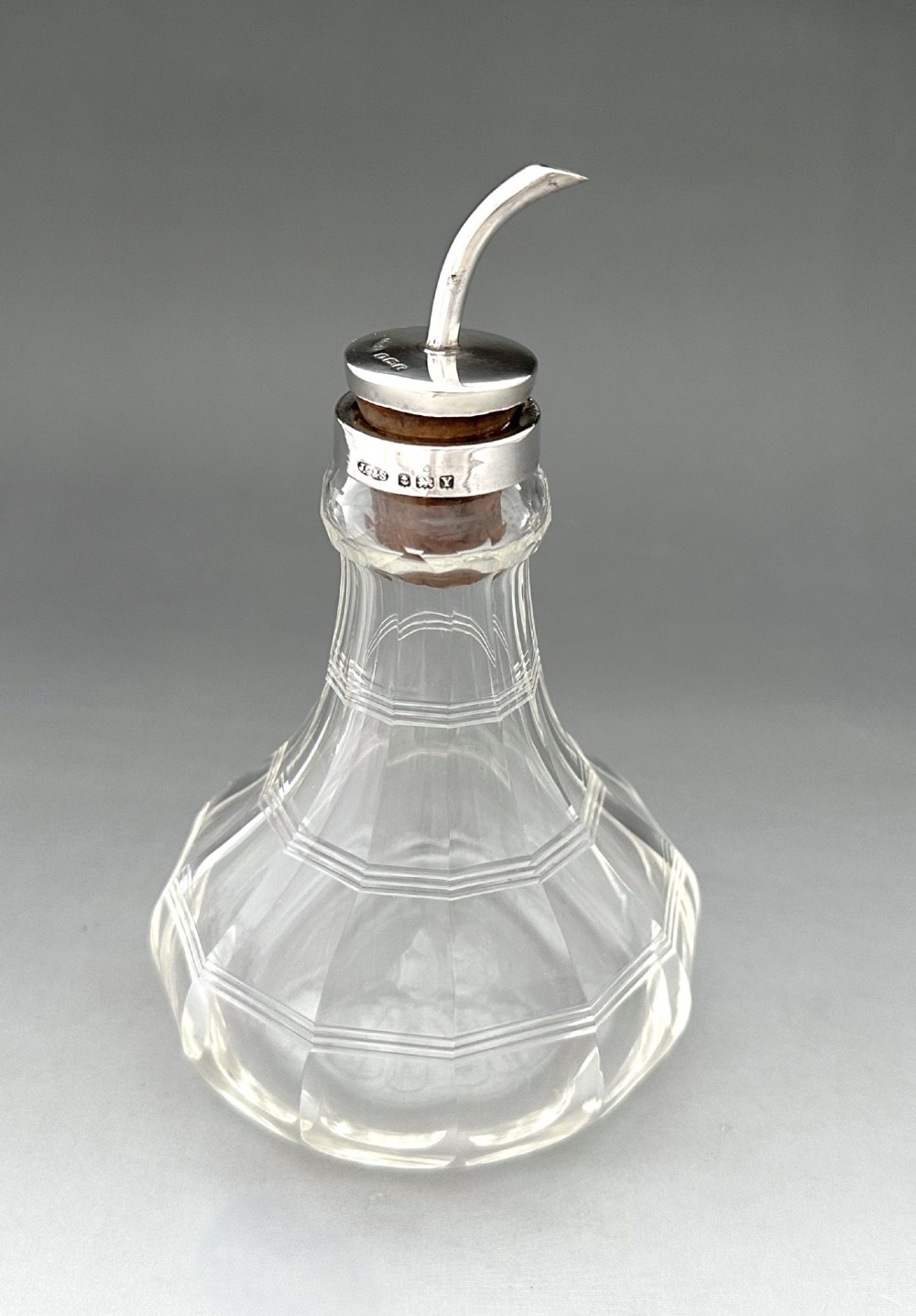 An Art Deco clear faceted glass bitters bottle featuring a hallmarked silver collar and curved pouring spout stopper.