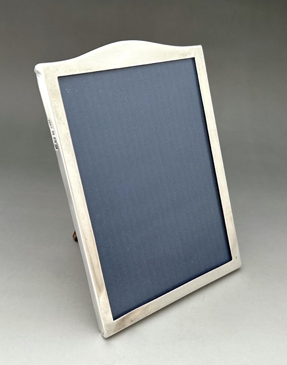 A rectangular sterling silver photo frame featuring a gently arched top edge, hallmark stamps along the side, and a dark blue insert.