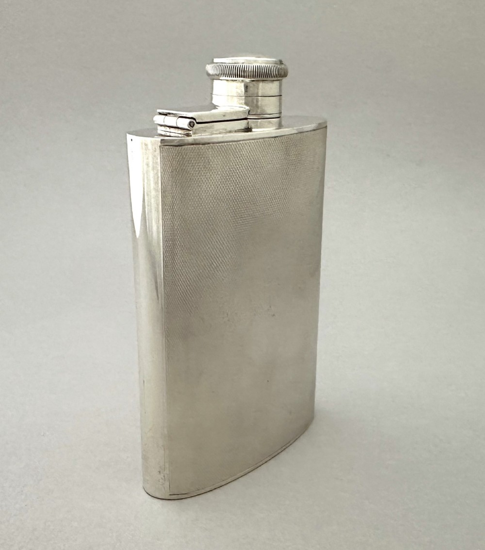 An Art Deco silver curved hip flask featuring fine engine-turned geometric detailing and a hinged cap. View II