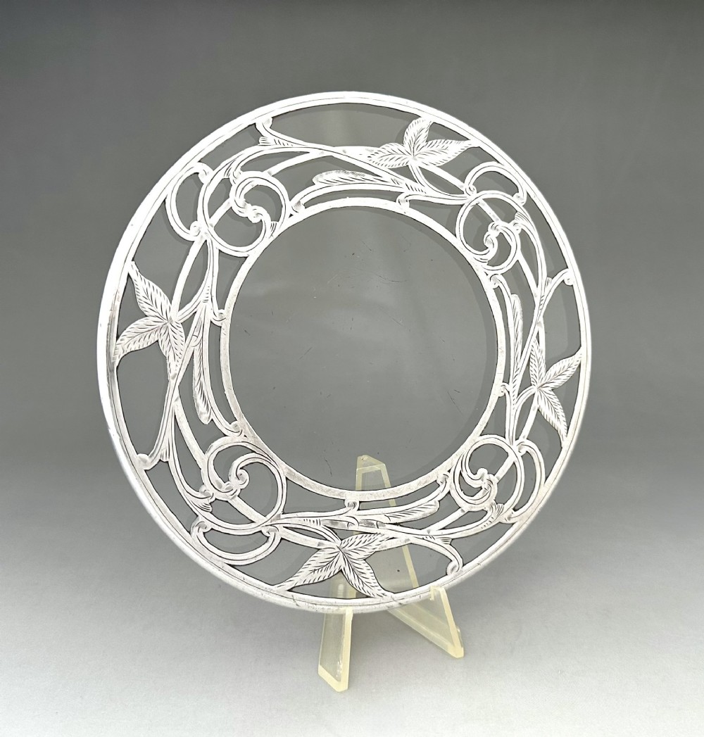 A circular clear glass teapot stand featuring a sterling silver rim with openwork Art Nouveau leaf motifs.