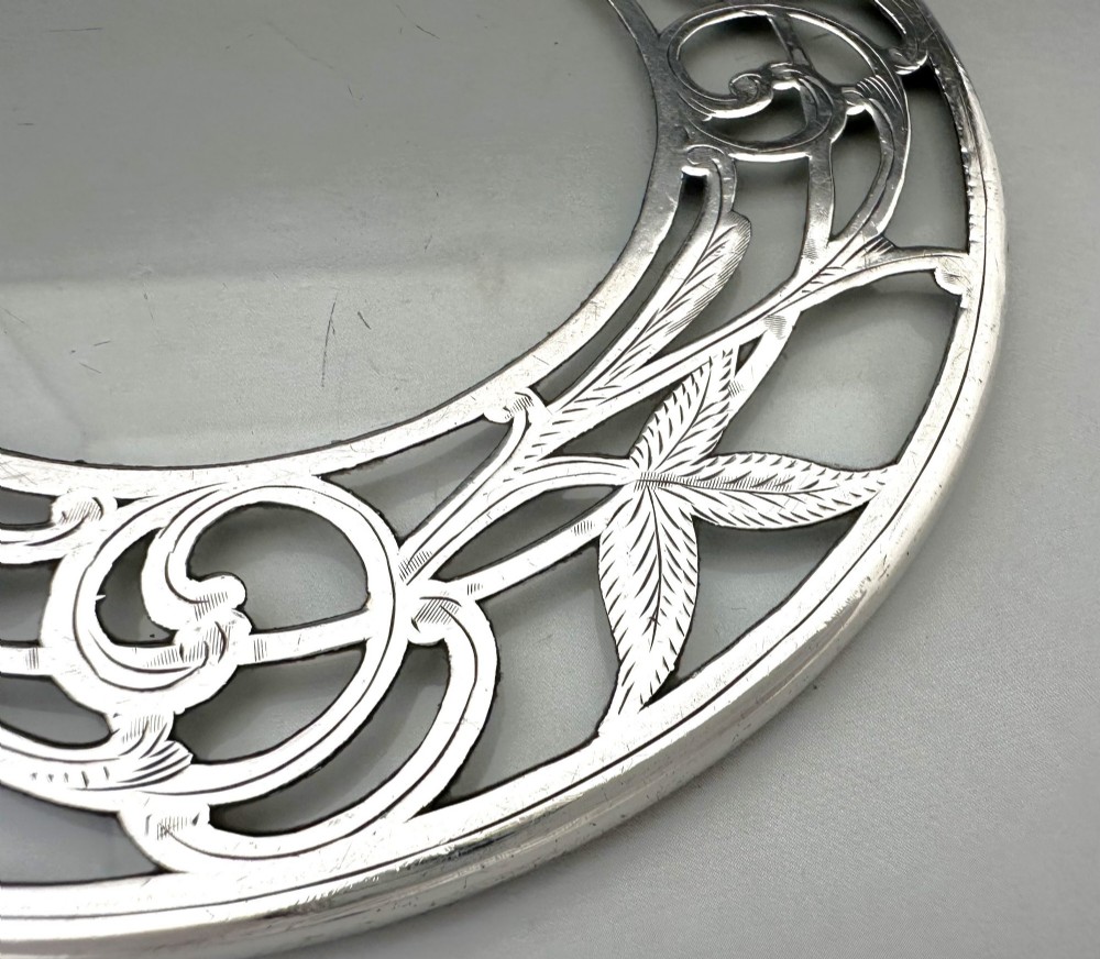 A circular clear glass teapot stand featuring a sterling silver rim with openwork Art Nouveau leaf motifs. View IV