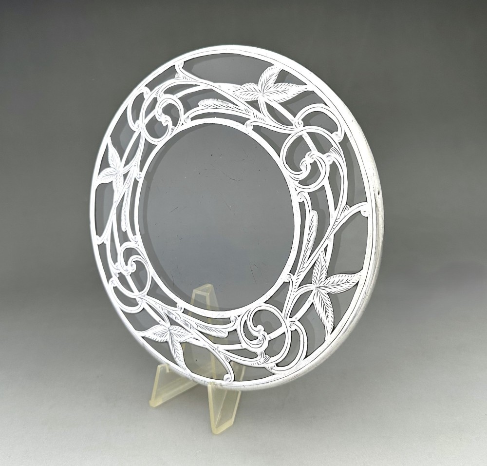 A circular clear glass teapot stand featuring a sterling silver rim with openwork Art Nouveau leaf motifs. View V