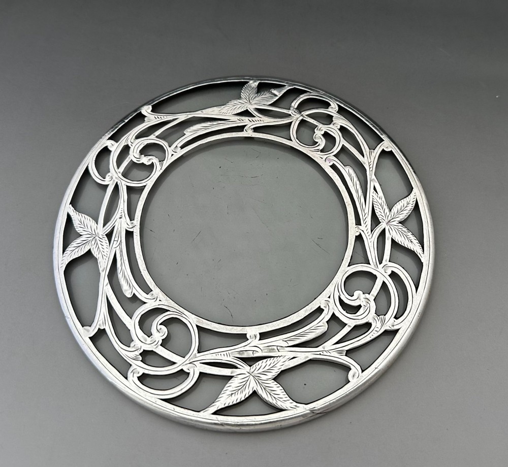 A circular clear glass teapot stand featuring a sterling silver rim with openwork Art Nouveau leaf motifs. View VI
