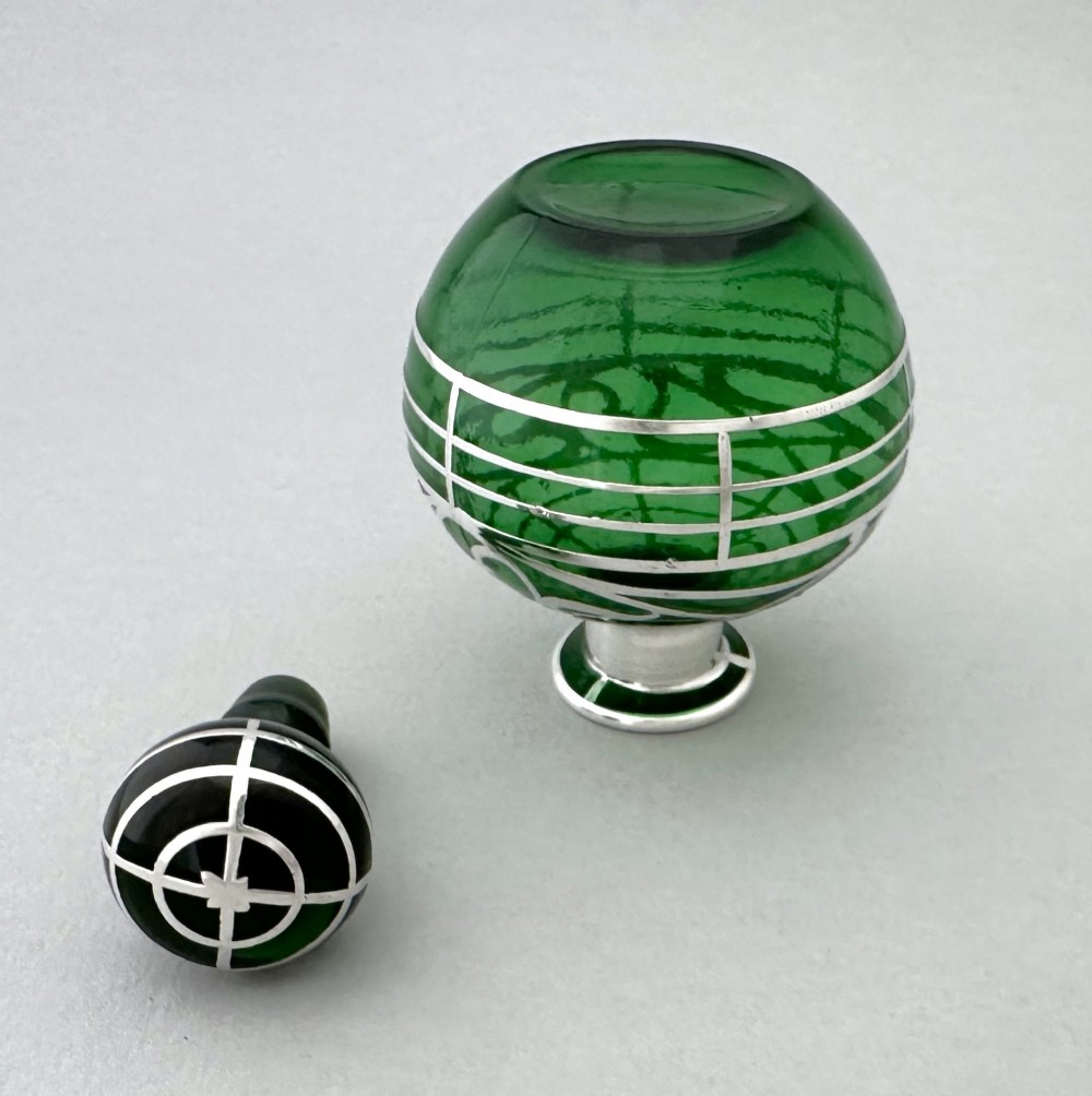 A globular green glass scent bottle featuring intricate silver overlay scrollwork and a matching fitted stopper. View V