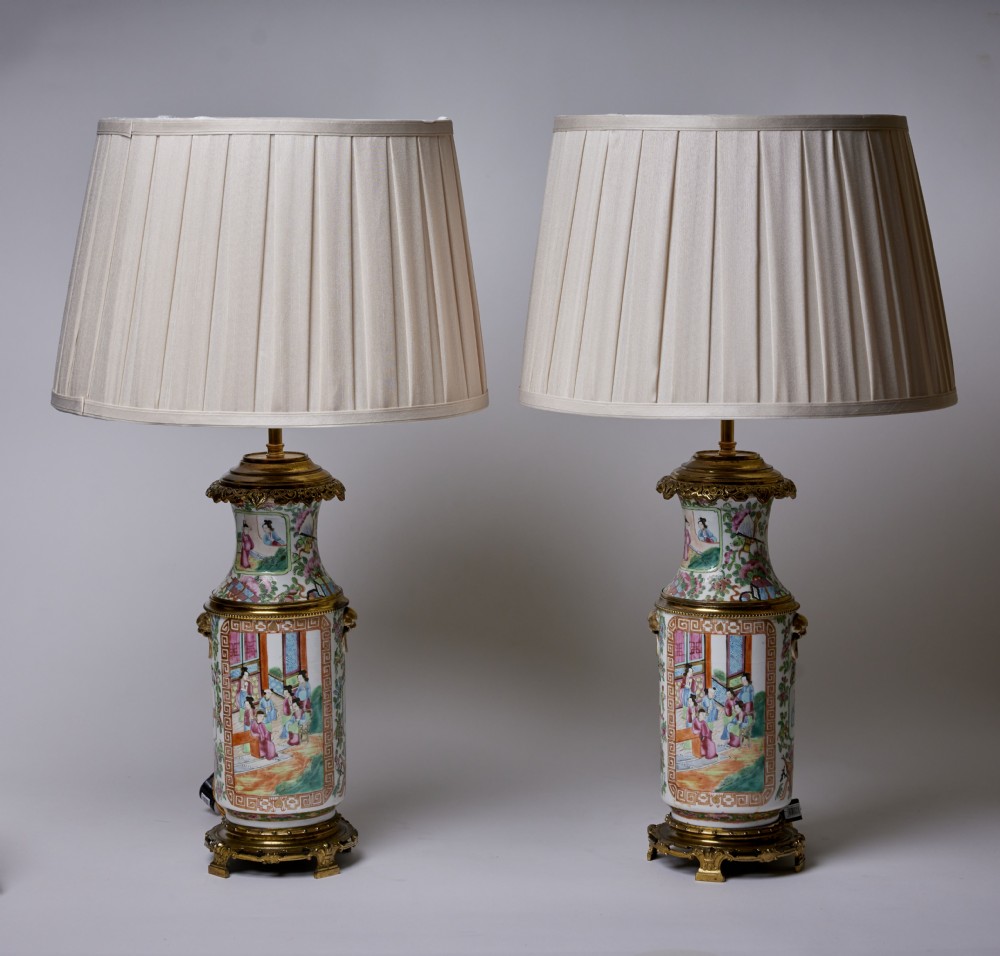 A pair of Chinese porcelain vase table lamps with hand-painted figures, gilt metal mounts, and cream pleated fabric shades.