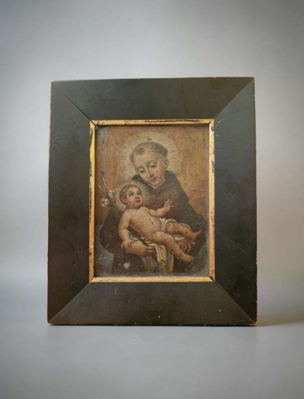 An antique Italian devotional icon depicting a saint holding the Christ Child set within a wide ebonised wooden frame with a gilded inner moulding.