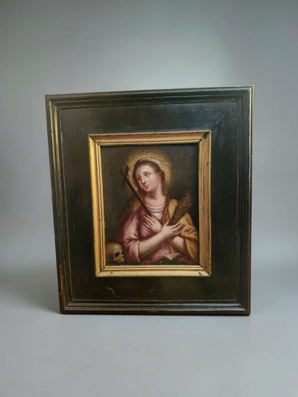 A small Flemish style religious oil painting depicting a female saint holding a cross and palm frond, housed in a thick ebonised wood frame with a gold sight edge.