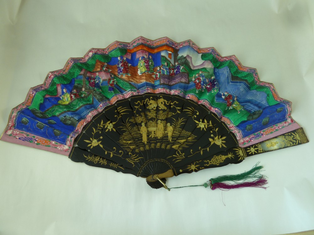 An open Chinese export folding hand fan displaying gilded black lacquer sticks and a paper leaf decorated with painted figures and colourful landscape borders.