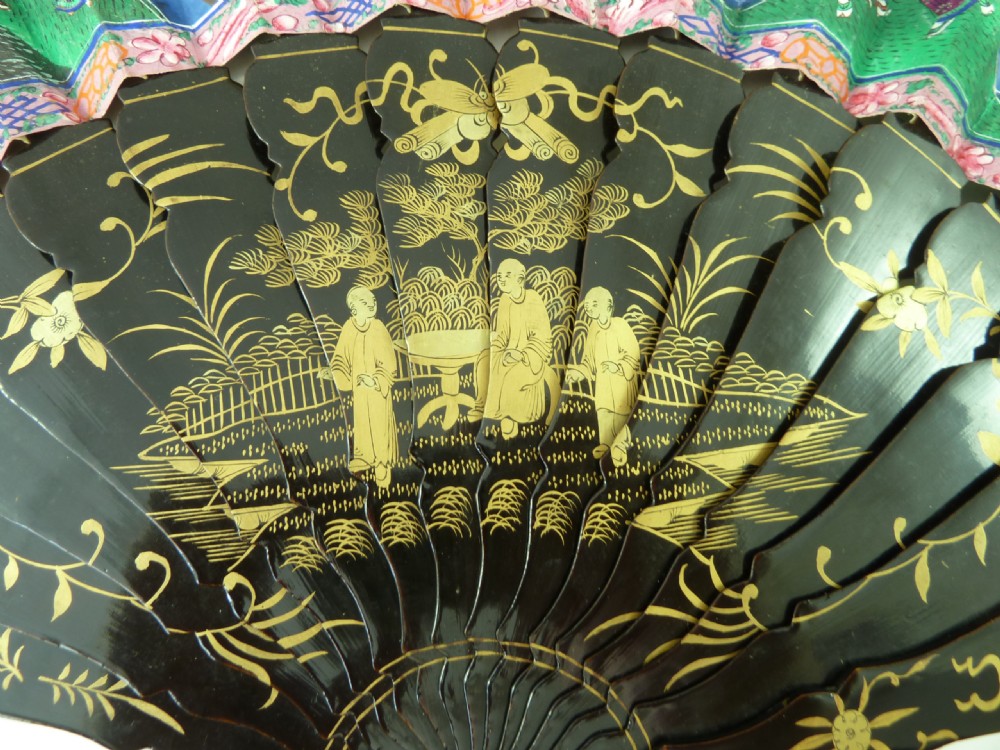 An open Chinese export folding hand fan displaying gilded black lacquer sticks and a paper leaf decorated with painted figures and colourful landscape borders. View II