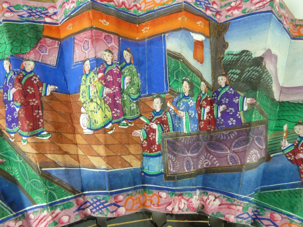 An open Chinese export folding hand fan displaying gilded black lacquer sticks and a paper leaf decorated with painted figures and colourful landscape borders. View III