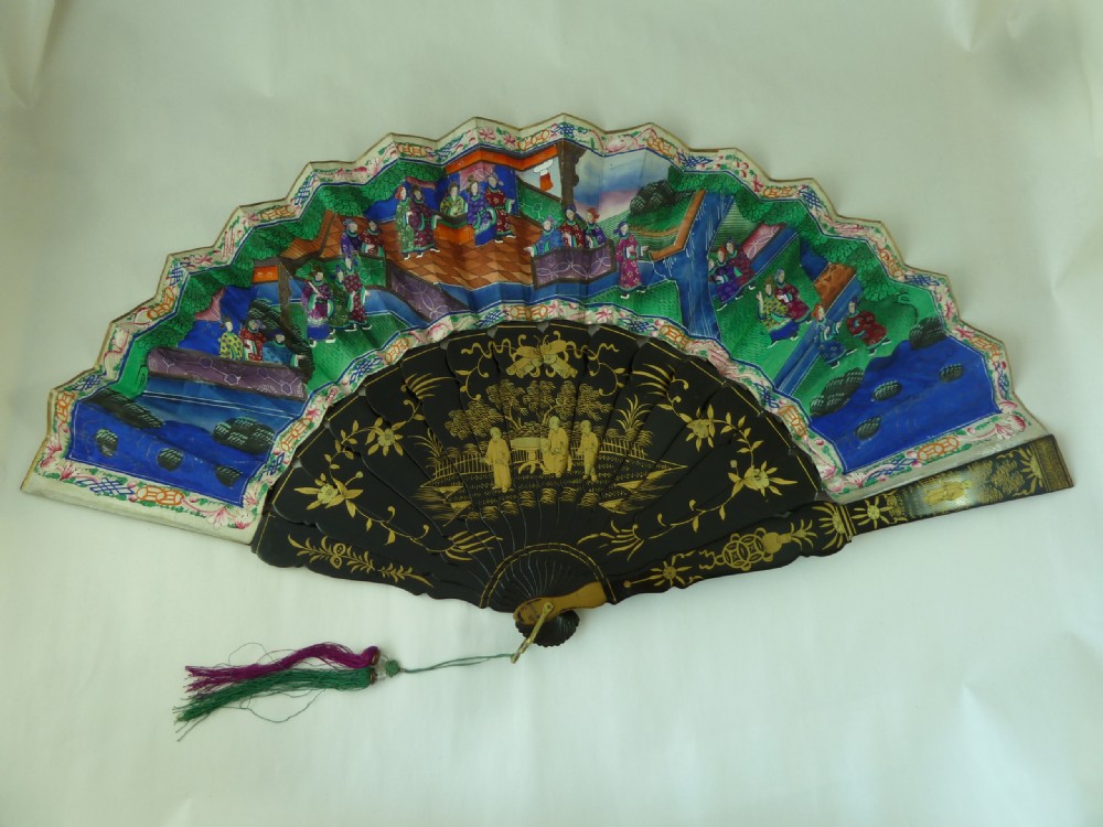 An open Chinese export folding hand fan displaying gilded black lacquer sticks and a paper leaf decorated with painted figures and colourful landscape borders. View IV
