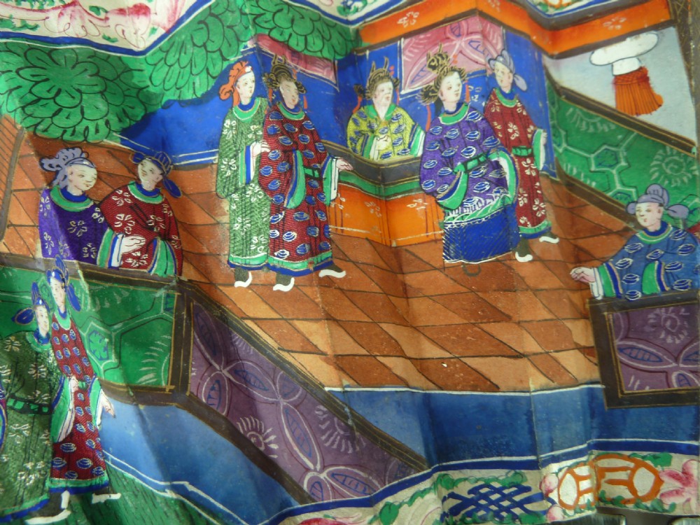 An open Chinese export folding hand fan displaying gilded black lacquer sticks and a paper leaf decorated with painted figures and colourful landscape borders. View V