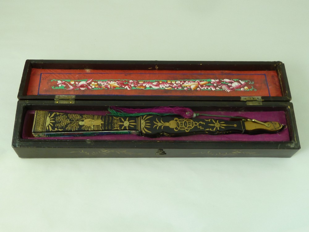 An open Chinese export folding hand fan displaying gilded black lacquer sticks and a paper leaf decorated with painted figures and colourful landscape borders. View VI
