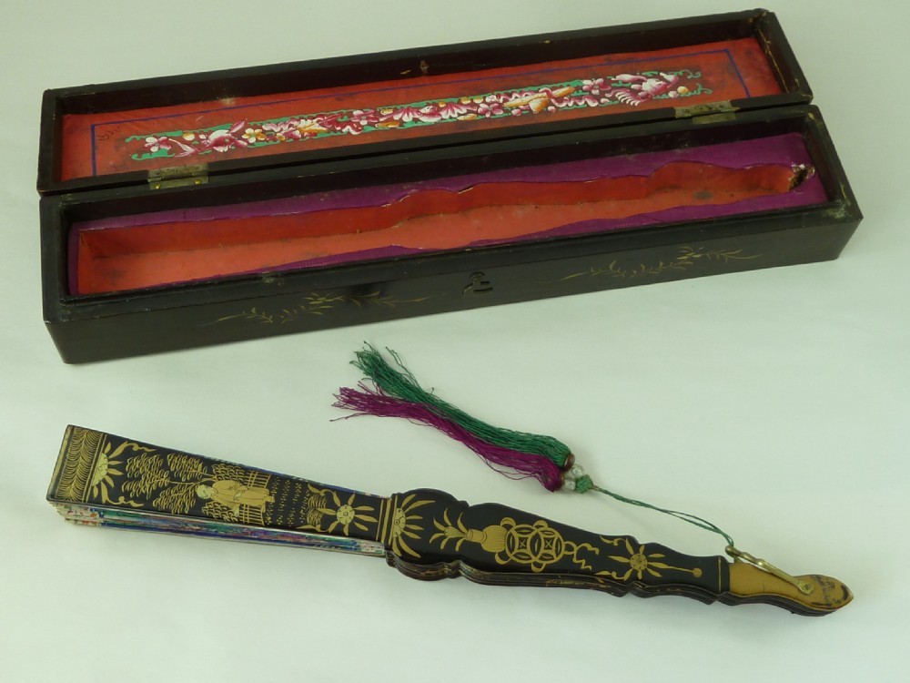 An open Chinese export folding hand fan displaying gilded black lacquer sticks and a paper leaf decorated with painted figures and colourful landscape borders. View VII