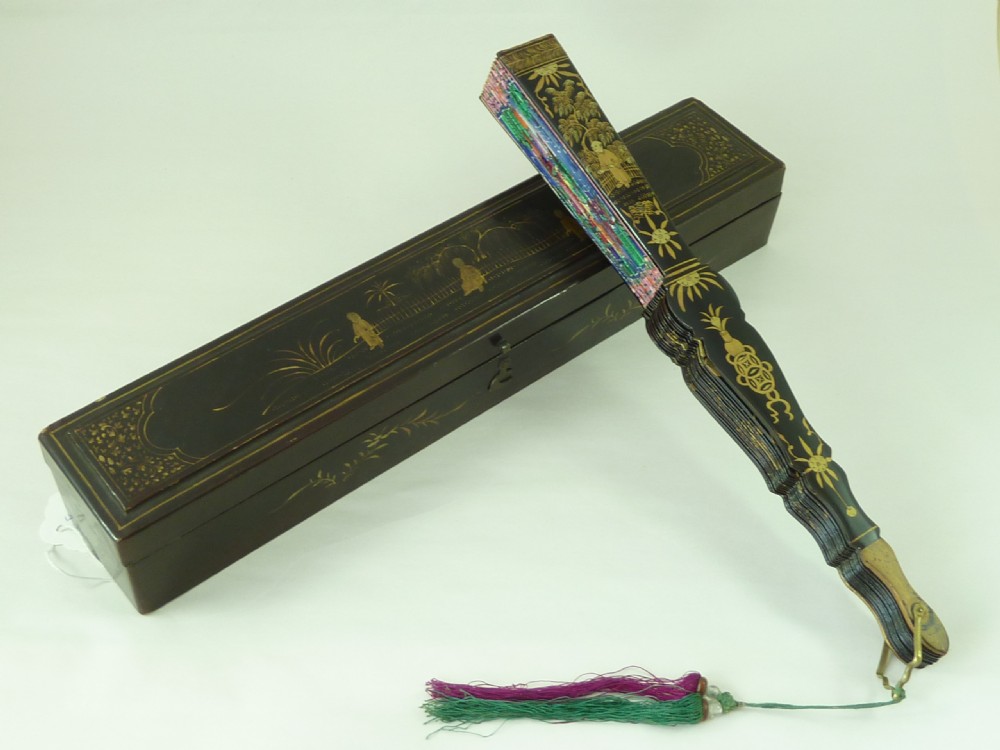 An open Chinese export folding hand fan displaying gilded black lacquer sticks and a paper leaf decorated with painted figures and colourful landscape borders. View VIII