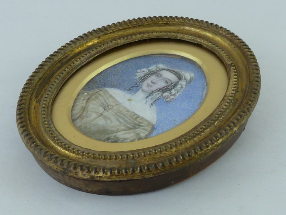 An oval 1830s portrait miniature painting depicting a lady with corkscrew ringlets and a lace cap against a blue background, contained in a gadrooned gilt brass frame. View II