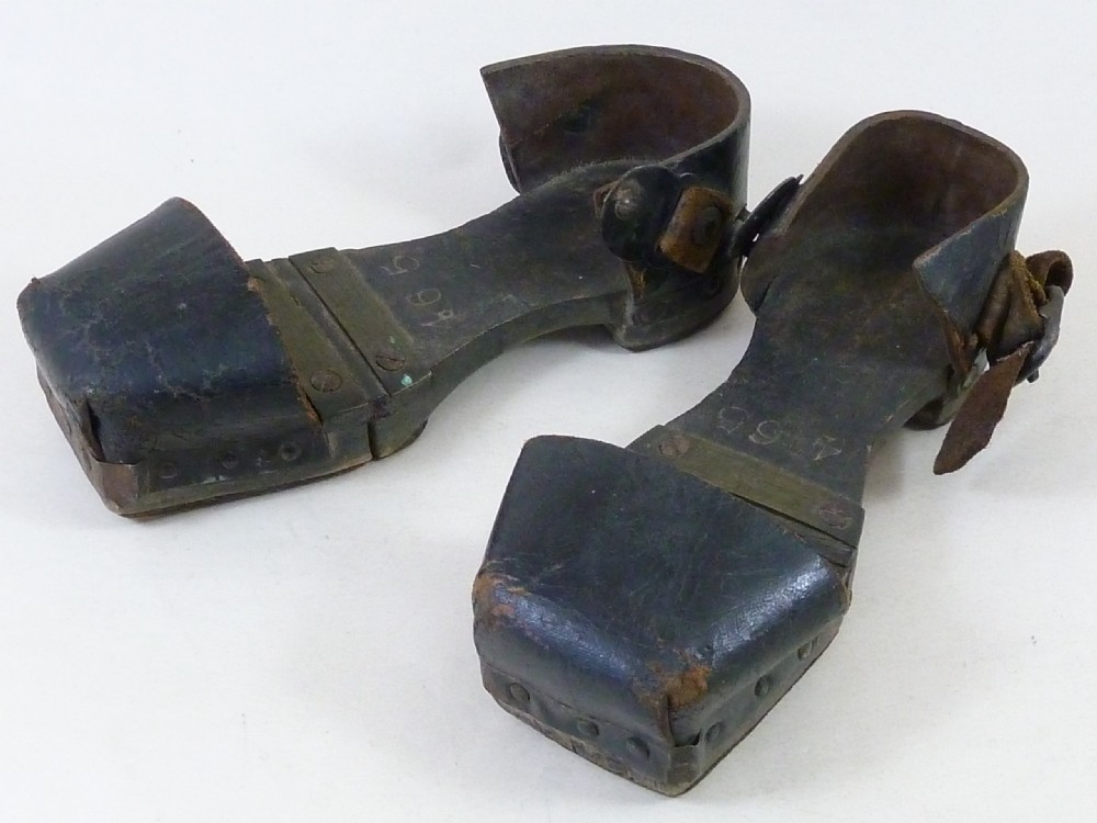 A pair of antique child hinged wooden overshoes featuring dark leather toe caps and heel straps with metal fittings.