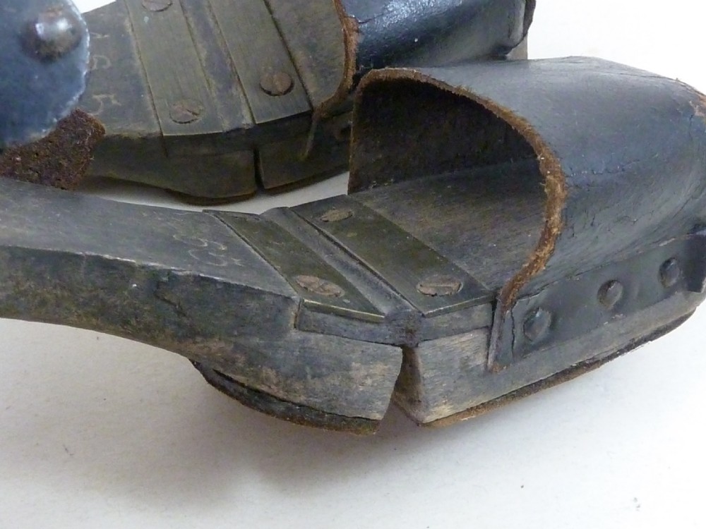 A pair of antique child hinged wooden overshoes featuring dark leather toe caps and heel straps with metal fittings. View V
