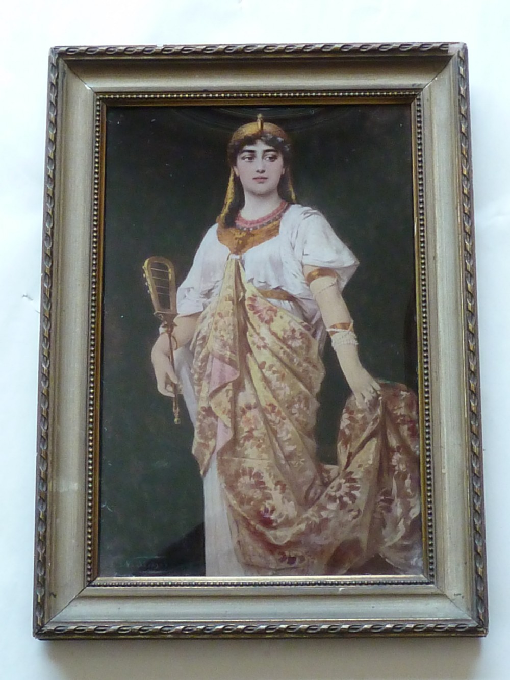 A framed Victorian chrystoleum reverse glass print depicting a figure in Egyptian Revival attire holding a musical instrument inside a gilded wooden frame.