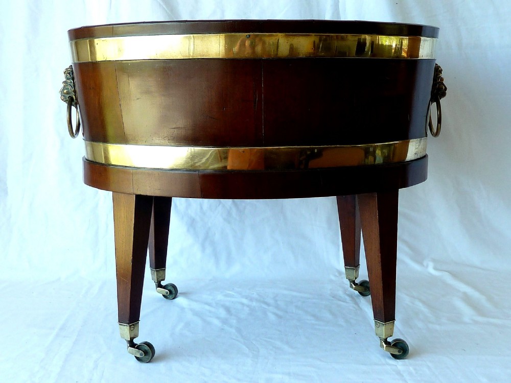 An oval Georgian mahogany wine cooler bound with two polished brass hoops, featuring lion mask ring handles and four tapered square legs with brass castors. View V