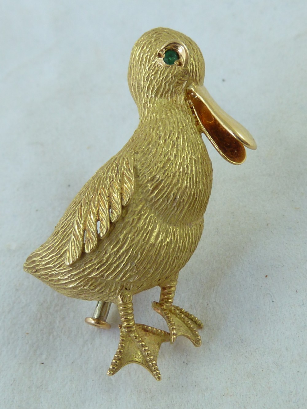 An 18ct yellow gold brooch designed as a standing duckling with textured feathers, an open bill, webbed feet, and a green emerald eye. View VIII