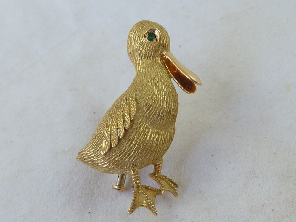 An 18ct yellow gold brooch designed as a standing duckling with textured feathers, an open bill, webbed feet, and a green emerald eye. View IX