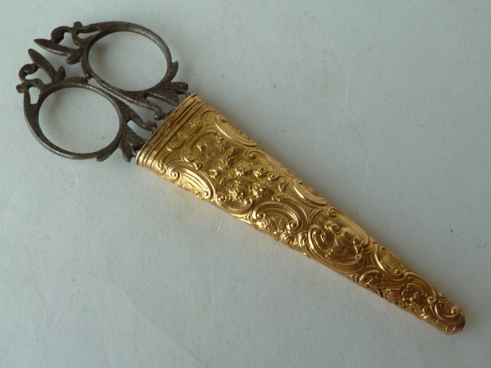 An 18th-century style tapered gold-coloured metal sheath embossed with intricate scrollwork, containing cut steel embroidery scissors.