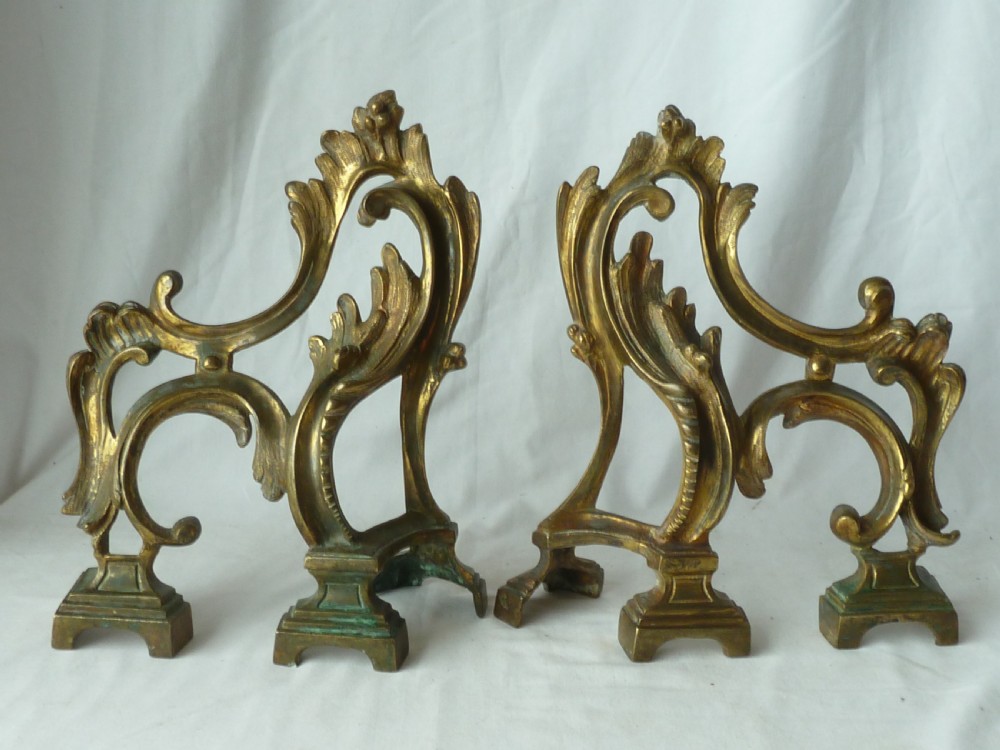 A pair of cast gilt brass French Rococo style fire dogs with elaborate acanthus leaf C-scroll design on small pedestal feet.