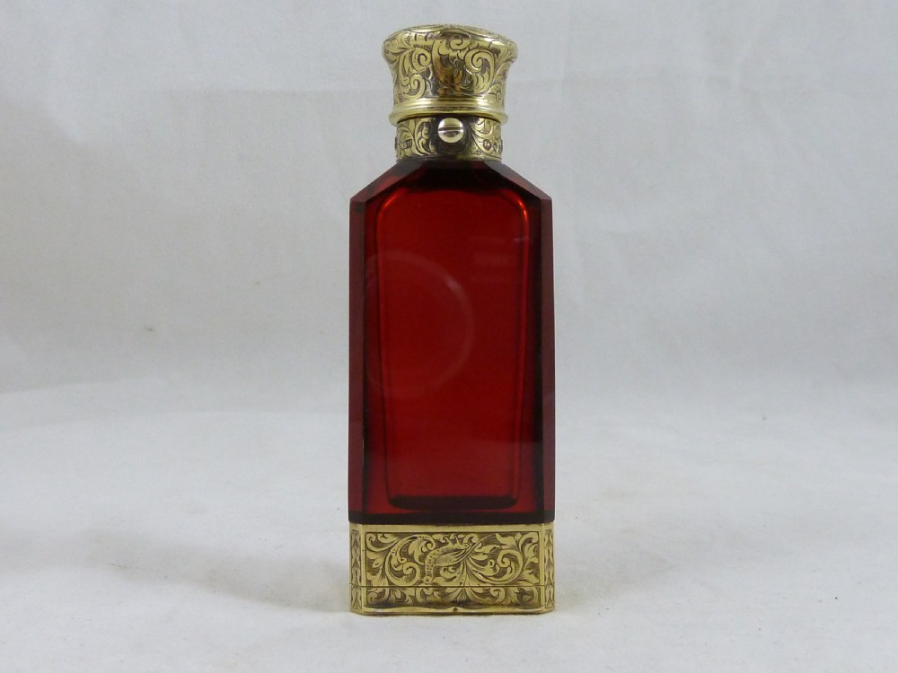 A faceted rectangular ruby red glass scent bottle featuring intricate scroll-engraved silver-gilt mounts at the cap and base.