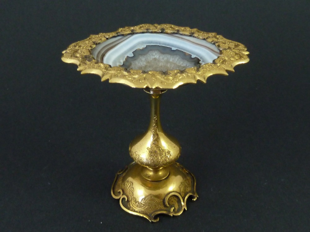 A miniature gilt brass tazza bowl featuring a central polished banded agate stone set within a chased Rococo scrollwork edge and pedestal foot.