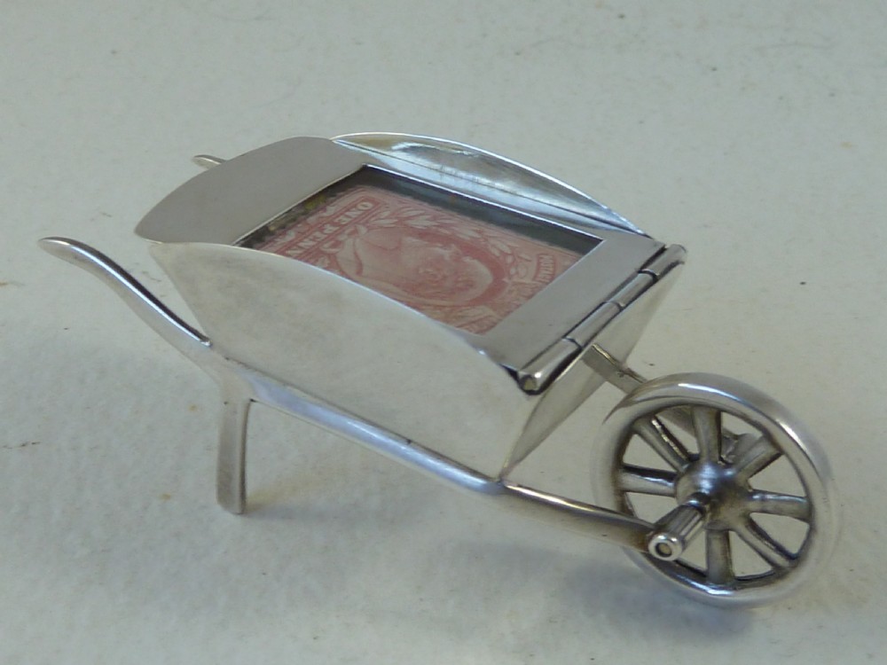 A miniature sterling silver stamp case shaped like a garden wheelbarrow with a clear glass window displaying a pink postage stamp.