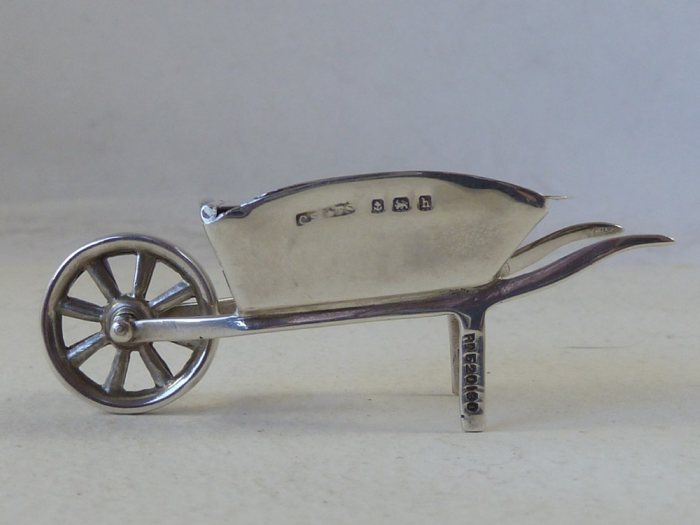 A miniature sterling silver stamp case shaped like a garden wheelbarrow with a clear glass window displaying a pink postage stamp. View II